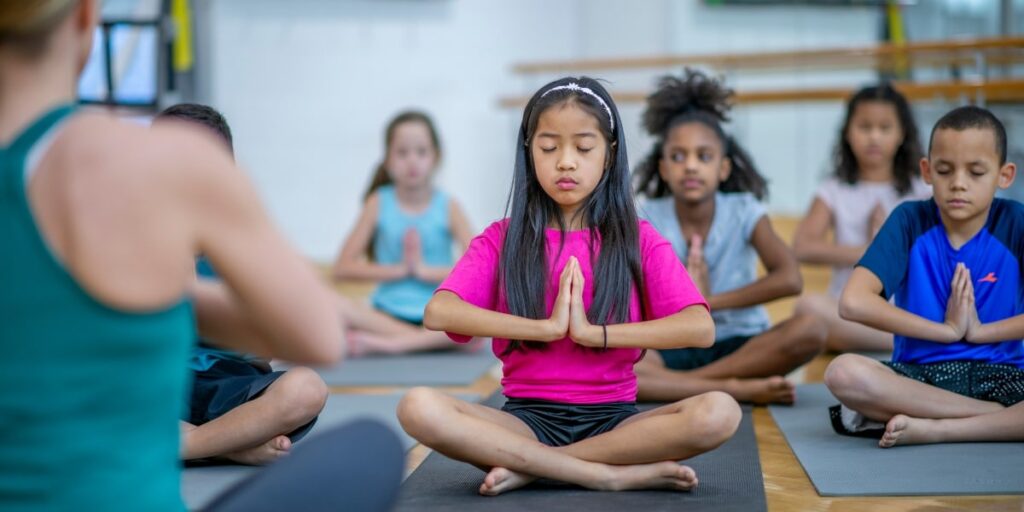girl with pink shirt meditating and practicing mindfulness, surrounded by other children.