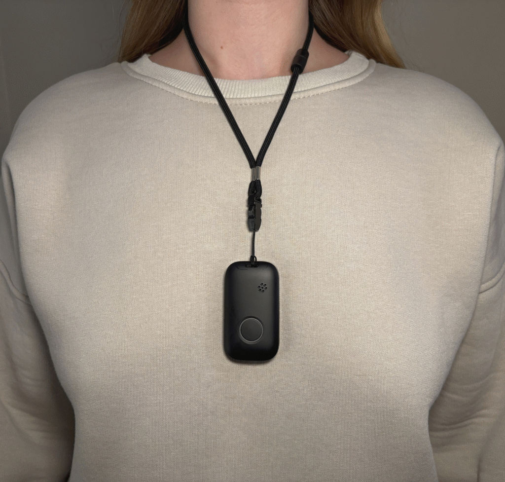 Black mobile alert device with a round button worn as a pendant on an adjustable black cord over a cream sweatshirt