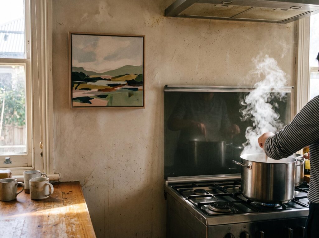 A framed canvas hangs on the far wall of a kitchen, away from a steaming pot and a sunny window.